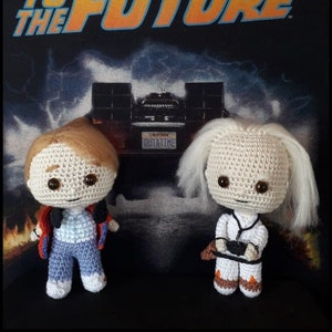 May include: Two crocheted figures from the movie Back to the Future. The figures are standing in front of a black t-shirt with the movie's logo. The figure on the left is wearing a red jacket, blue jeans, and white sneakers. The figure on the right is wearing a white jumpsuit, brown boots, and a white lab coat. The figures are both smiling.