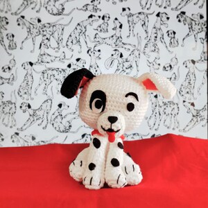 Op de afbeelding: Een wit en zwart gehaakt Dalmati&euml;r puppy speelgoed met rode oren en een rode tong die uitsteekt. Het speelgoed zit op een rood oppervlak.