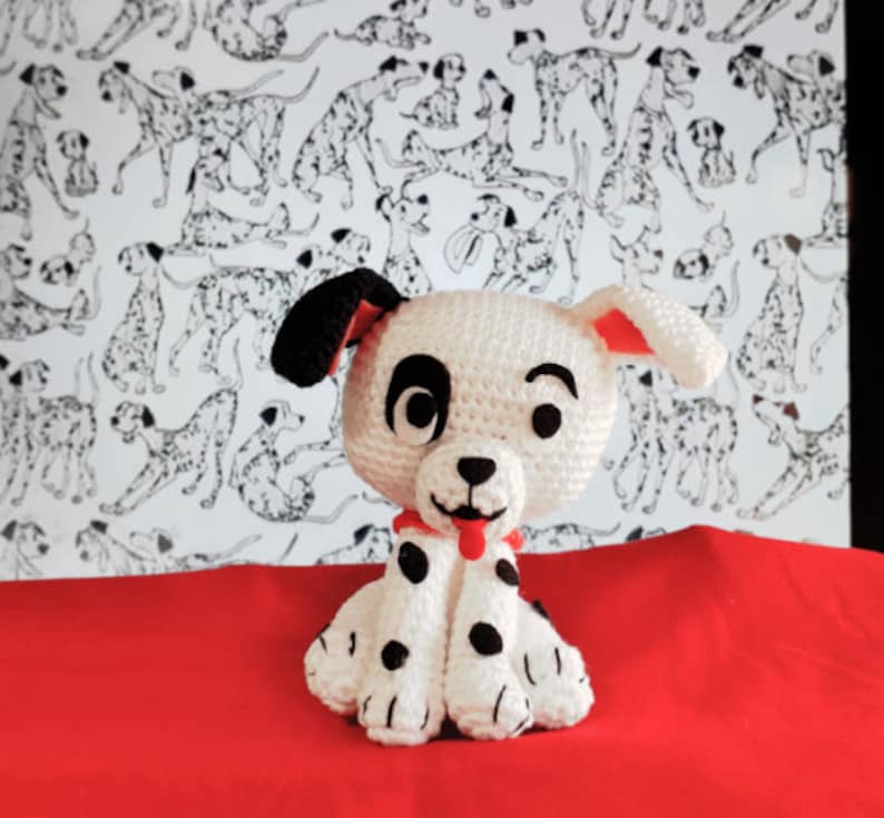 Op de afbeelding: Een wit en zwart gehaakt Dalmati&euml;r puppy speelgoed met rode oren en een rode tong die uitsteekt. Het speelgoed zit op een rood oppervlak.