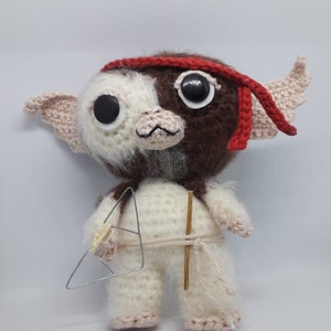 Può includere: Un giocattolo di peluche all'uncinetto di un personaggio con pelliccia marrone e bianca, che indossa una fascia rossa e tiene un bastone di legno e un triangolo di filo. Il giocattolo ha grandi occhi neri e un naso rosa.