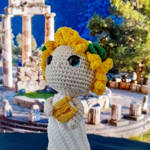 Puede incluir: Una muñeca de crochet con cabello amarillo, un vestido blanco y una lira amarilla. La muñeca está de pie frente a un fondo borroso de un antiguo templo griego.