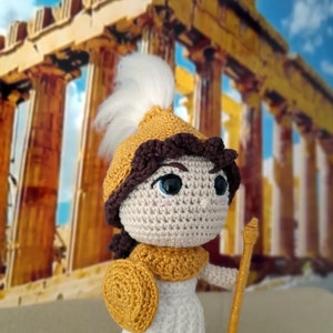Könnte beinhalten: Eine gehäkelte Puppe, die ein weißes Kleid, einen gelben Helm mit einer weißen Feder und einen gelben Schild trägt. Die Puppe hält eine gelbe Lanze und steht vor einem verschwommenen Bild eines steinernen Gebäudes.