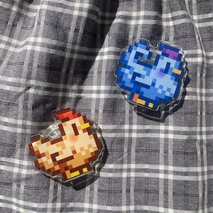 May include: Two pixel art-style acrylic pins. One pin features a brown and yellow chicken, and the other a blue bird. The pins are on a gray and white plaid fabric background.