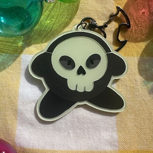 May include: A green and black key chain charm featuring a cartoon skull with a black outline. The charm is attached to a black metal ring.
