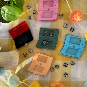 Puede incluir: Cinco estuches de juegos de Nintendo DS de plástico de colores vivos con una variedad de cartuchos de juegos. Los estuches están dispuestos sobre una superficie a cuadros amarilla y blanca.