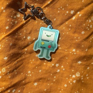 May include: A light blue keychain featuring a cartoon character with a green face and a red button. The keychain has a silver star-shaped clasp and a silver keyring. The background is a textured orange fabric with white star and flower patterns.