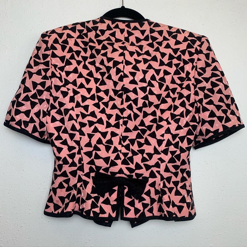 May include: A pink and black short-sleeved blouse with a bow at the neckline. The blouse is made of a silky fabric and has a pattern of black bow ties on a pink background.