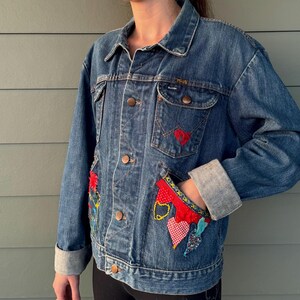 Könnte beinhalten: Eine Vintage-Jeansjacke mit klassischem Kragen, Knopfverschlüssen und Taschen. Die Jacke hat farbenfrohe Stoffflicken mit Herz- und Blumenmotiven auf den Taschen und Ärmeln. Die Manschetten sind hochgekrempelt und zeigen ein helleres Jeansfutter.
