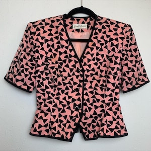 May include: A pink and black short-sleeved blazer with a bow tie pattern. The blazer has a button-up front and a fitted silhouette. The label reads "Nicole Miller".