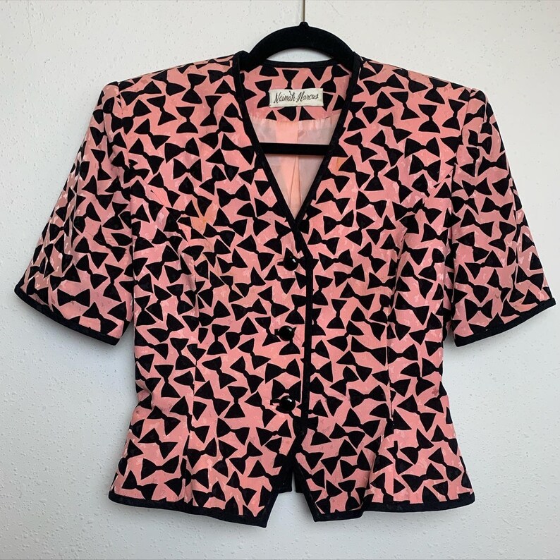 May include: A pink and black short-sleeved blazer with a bow tie pattern. The blazer has a button-up front and a fitted silhouette. The label reads "Nicole Miller".