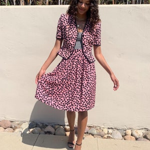 May include: A pink and black patterned skirt suit with a short-sleeved jacket and a knee-length skirt. The skirt has a pleated design and the jacket has a bow detail at the neckline.