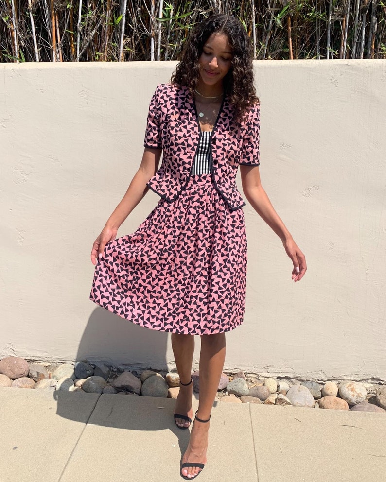 May include: A pink and black patterned skirt suit with a short-sleeved jacket and a knee-length skirt. The skirt has a pleated design and the jacket has a bow detail at the neckline.