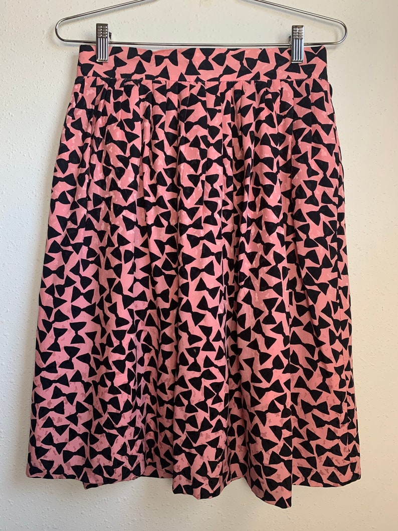 May include: A pink midi skirt with a black bow print. The skirt has a pleated design and a high waist.