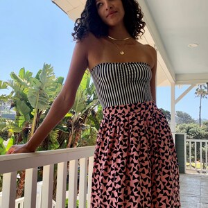 May include: A woman wearing a black and white striped strapless top and a pink skirt with a black geometric pattern. She is standing on a balcony with a white railing and looking up.