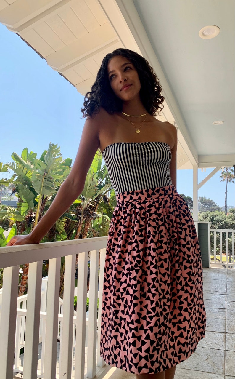 May include: A woman wearing a black and white striped strapless top and a pink skirt with a black geometric pattern. She is standing on a balcony with a white railing and looking up.