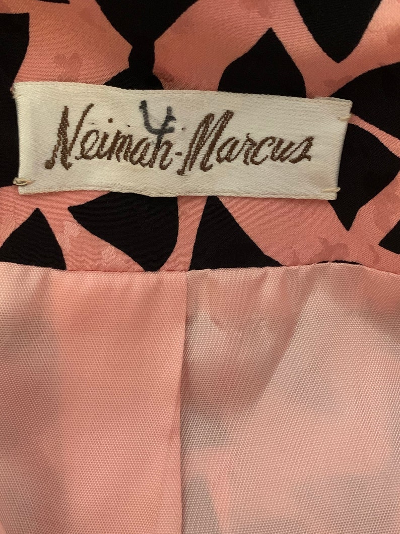 May include: A white label with the text "Neiman-Marcus" in brown lettering. The label is attached to a pink and black fabric with a geometric pattern.