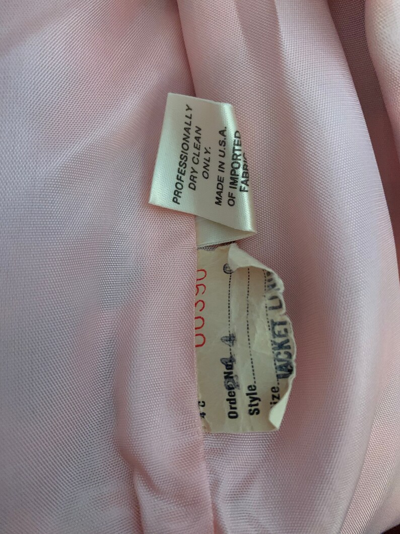 May include: Close-up of a pink fabric label with the text "Professionally Dry Clean Only. Made in U.S.A. Of Imported Fabric" and a white label with the text "Order No. 4 Style. Size Jacket Lining 00390 4C".