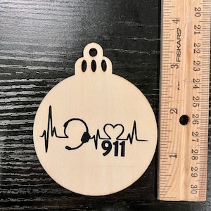May include: A wooden ornament with a black graphic of a heartbeat, a headset, a heart, and the number 911. The ornament is round with a decorative top. A wooden ruler is next to the ornament.