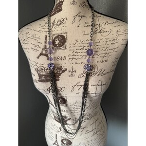 May include: A long, layered necklace with a gunmetal finish. The necklace features strands of chain, purple beads, and a floral pendant with purple accents. The necklace is displayed on a mannequin with vintage-style script.