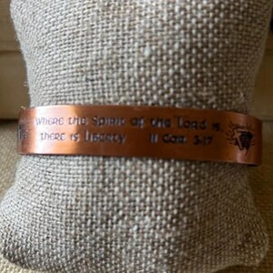 May include: A copper cuff bracelet featuring the engraved text "Where the Spirit of the Lord is, there is liberty" followed by "II Cor. 3:17" and a small cross symbol. The metal has a polished finish with dark, recessed lettering.