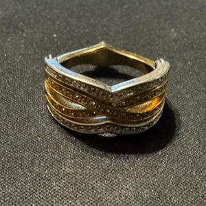 May include: A two-tone ring featuring a V-shaped design with alternating bands of silver-colored metal and gold-colored metal. The silver-colored sections are encrusted with small, clear, sparkling stones, while the gold-colored bands are smooth.
