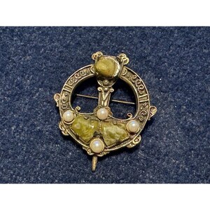 May include: A circular metal brooch featuring an intricate engraved pattern along the outer ring. The piece is adorned with three small white pearls and three faceted olive-green stones, with a central pin mechanism visible across the back.