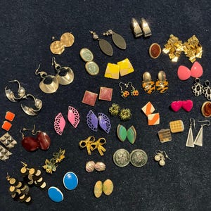 May include: A collection of assorted earrings including gold-tone hoops, geometric shapes, floral designs, and colorful studs. The set features various materials and styles, such as dangling metal pieces, bright pink hearts, blue ovals, and textured squares.