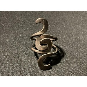 May include: A dark metallic ring featuring a continuous, swirling design that wraps around the finger. The band is constructed from a single piece of metal that curves into multiple loops, creating an open, fluid, and interconnected spiral pattern.
