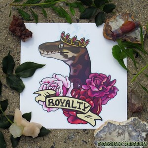 May include: A digital illustration of a snake wearing a gold crown with red jewels. The snake is surrounded by pink and red roses and a banner that reads "ROYALTY".