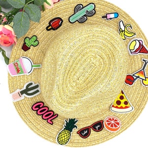 May include: A straw hat adorned with colorful embroidered patches. Patches include a cactus, ice cream, pizza slice, pineapple, and sunglasses with the text "hello you". The hat is surrounded by pink roses and green leaves.