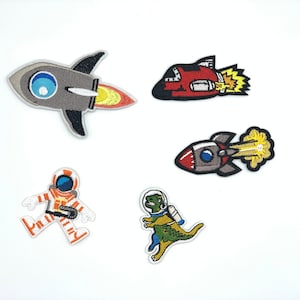 May include: Five embroidered iron-on patches featuring a space theme. The patches include a gray rocket, a red rocket, a gray rocket with a blue flame, a white astronaut, and a green dinosaur wearing a helmet.