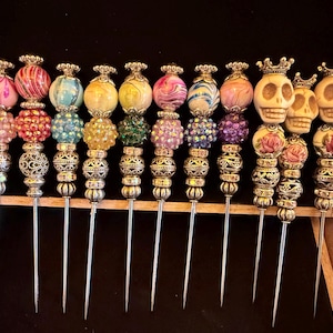May include: A collection of eleven decorative metal skewers featuring ornate silver-toned spacers and colorful beads. Eight skewers are topped with marbled or faceted glass spheres in various colors, while three feature carved skull beads with crowns.
