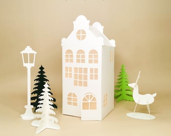 May include: White papercraft model of a European-style house with a deer, two Christmas trees, and a street lamp. The house has a white roof and windows with a light brown interior. The deer is white with a brown horn. The trees are green. The street lamp is white with a black top.