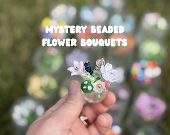 Op de afbeelding: Een miniatuurboeket van kralenbloemen in een klein doorzichtig glazen vaasje, met roze, witte en groene glaskralen in de vorm van bloemblaadjes en een groene paddenstoelkraal met witte stippen. De tekst MYSTERY BEADED FLOWER BOUQUETS staat erboven.
