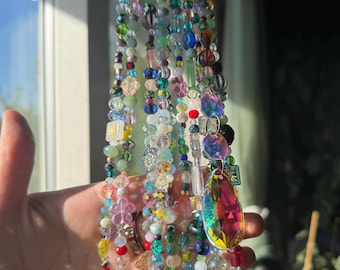 Op de afbeelding: Een kleurrijke kralen zonnevanger met kralen van verschillende vormen en maten in tinten blauw, groen, roze en geel. Een groot, iriserend, druppelvormig kristal hangt onderaan.
