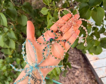 Op de afbeelding: Handgemaakte kralen spinnenornamenten in blauw, groen en roze. Elke spin heeft delicaat kralenwerk, goudkleurige draad en bungelende kristallen accenten. De spinnen worden getoond tegen een achtergrond van groen gebladerte, wat hun ingewikkelde ontwerpen laat zien.