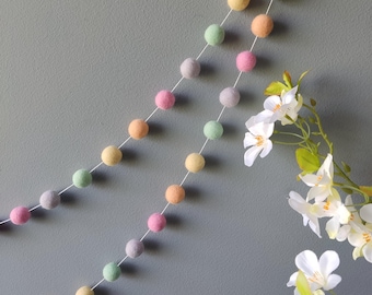 Puede incluir: Una guirnalda de bolas de fieltro de color pastel con una cuerda blanca. La guirnalda está colgada en una pared gris y tiene una variedad de colores, incluyendo rosa, verde, amarillo y azul claro.