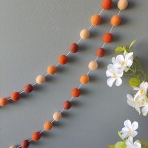 May include: A garland of felt balls in shades of orange, brown, and beige. The garland is strung on a white string and is hanging against a grey wall.