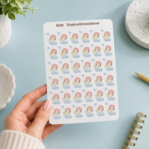 May include: A white rectangular sticker sheet featuring thirty-five small illustrations of a character in a pink shower cap sitting in a blue bubble bath. The text Bath Empirestickerplanner is printed in brown at the top of the sheet.