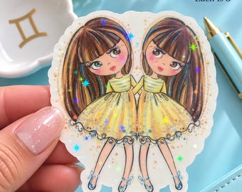 May include: A die-cut sticker featuring two identical cartoon characters with long brown hair, wearing yellow dresses with striped tops and blue ballet slippers. The sticker has a holographic finish with star accents and measures 3 inches.