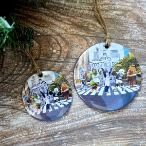 May include: Two round Christmas ornaments with a colorful image of characters crossing a street, resembling the iconic Abbey Road photo. The ornaments are hanging by twine strings, with evergreen branches in the background.