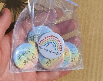May include: A white organza bag filled with colorful round buttons. The buttons have a rainbow design and the text "little bag of comfort".