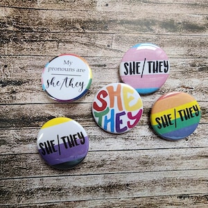May include: Five round buttons with different pride flag designs. Each button has the text "she/they" printed on it. One button has the text "My pronouns are she/they".
