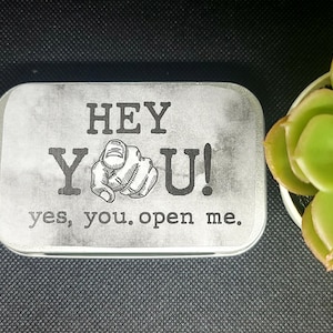 May include: A silver tin with a white background and black text that reads "HEY YOU! yes, you. open me.". The text is in a vintage style font. A black and white illustration of a hand pointing at the viewer is between the words "HEY" and "YOU!"