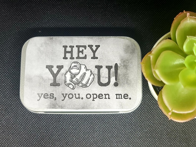 May include: A silver tin with a white background and black text that reads "HEY YOU! yes, you. open me.". The text is in a vintage style font. A black and white illustration of a hand pointing at the viewer is between the words "HEY" and "YOU!"