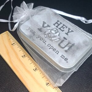May include: A small silver tin with a white sheer fabric pouch. The tin has the text "Hey You! Yes, you. Open me." printed on it.
