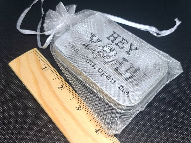 May include: A small silver tin with a white sheer fabric pouch. The tin has the text "Hey You! Yes, you. Open me." printed on it.