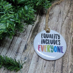 May include: White ceramic ornament with the text "Equality includes everyone" in rainbow colors. The ornament is hanging from a twine string.