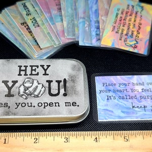 May include: A silver metal tin with the words "HEY YOU! yes, you. open me." printed on the front. The tin contains a set of colorful affirmation cards with inspirational messages. The cards are printed with a watercolor background.