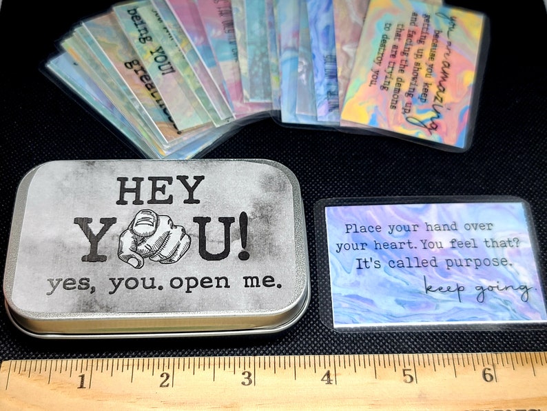 May include: A silver metal tin with the words "HEY YOU! yes, you. open me." printed on the front. The tin contains a set of colorful affirmation cards with inspirational messages. The cards are printed with a watercolor background.